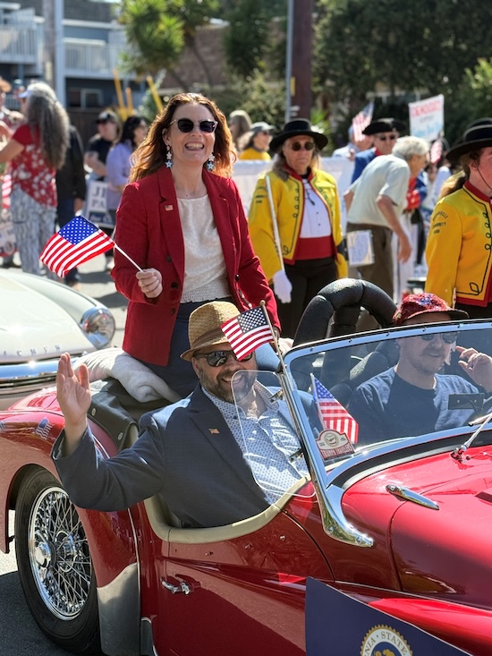 Asm Addis riding in the parade with my husband in a red car driven by a member of the Triumph Travelers Sports Car Club, waving an American flag
