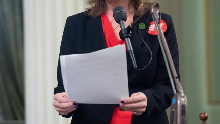 Asm. Addis speaking into microphone, holding papers