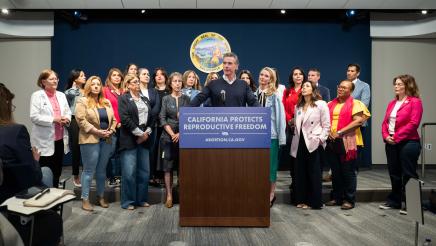 Go. Newsom at podium, speaking, with legislators standing in support