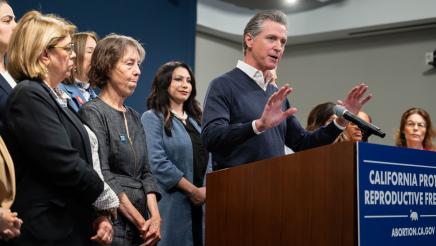 Go. Newsom at podium, speaking, with legislators standing in support