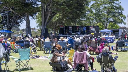 Large audience in grassy area listening to speaker onstage