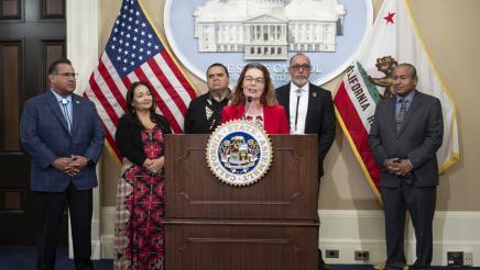 Asm. Addis at podium, speaking, with Asm. Ramos and guests in background