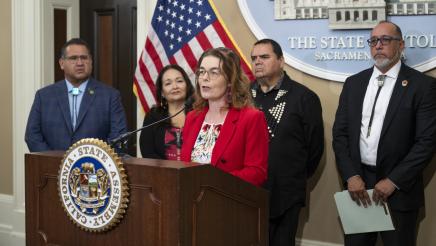 Asm. Addis at podium, speaking, with Asm. Ramos and guests in background