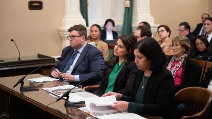 Witnesses, seated at desk, and audience members listening on
