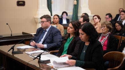 Witnesses, seated at desk, and audience members listening on