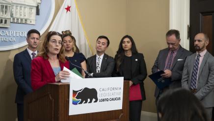 Asm. Addis at podium, speaking, while caucus members listen on