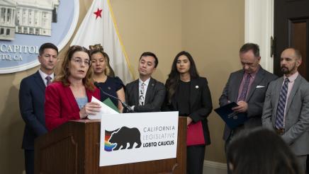 Asm. Addis at podium, speaking, while caucus members listen on