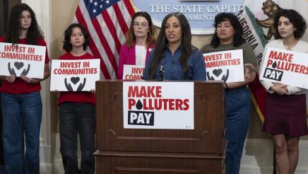 Jessica Calix at podium, speaking, as Asm. Addis and others listen on